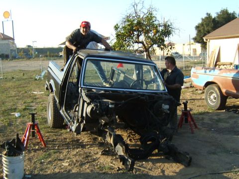 1988 Ford Bronco - Two men work on the disassembled chassis of a 1988 Ford Bronco, with another truck visible in the background.