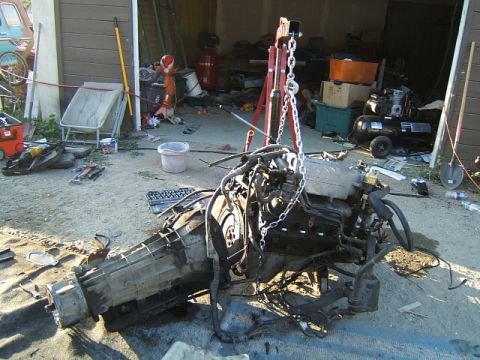 1988 Ford Bronco - An engine and transmission assembly is suspended by chains from an engine hoist, with various tools and equipment scattered around aorund t…