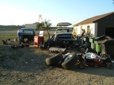 1988 Ford Bronco - A yard is filled with vehicle parts, two trucks, and a go-kart under a clear sky.