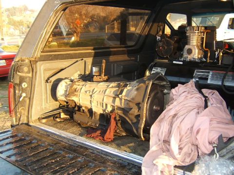 1988 Ford Bronco - A transmission and air compressor are loaded into the bed of a truck, with another vehicle visible in the background.