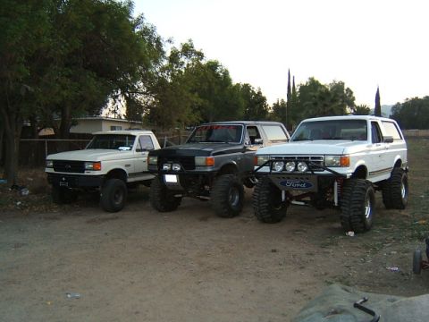 1988 Ford Bronco - Three modified Ford trucks and SUVs are parked on a dirt lot under a bright sky.
