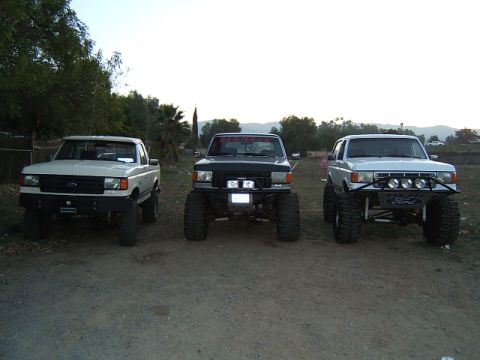 1988 Ford Bronco - Three lifted Ford trucks are parked side-by-side, viewed from the front. The middle truck displays red text across its windshield.