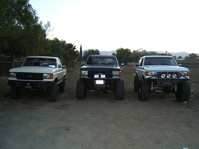 Three lifted Ford trucks in a row, front view. Middle truck has red text on windshield