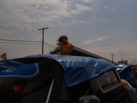 1993 Ford Bronco - A child sits atop a vehicle covered by a blue tarp with a Ford logo, under a cloudy sky.