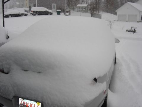 1999 Ford Taurus - A car is covered in a thick blanket of snow during a winter storm, with other vehicles and buildings visible.