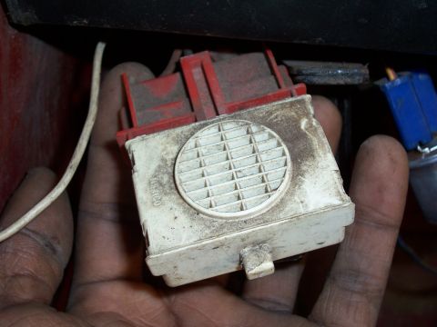 1978 Ford Bronco - Close-up of a hand holding a grimy white plastic buzzer/alarm module with a round grille face, unclipped from under the dash and still plug…