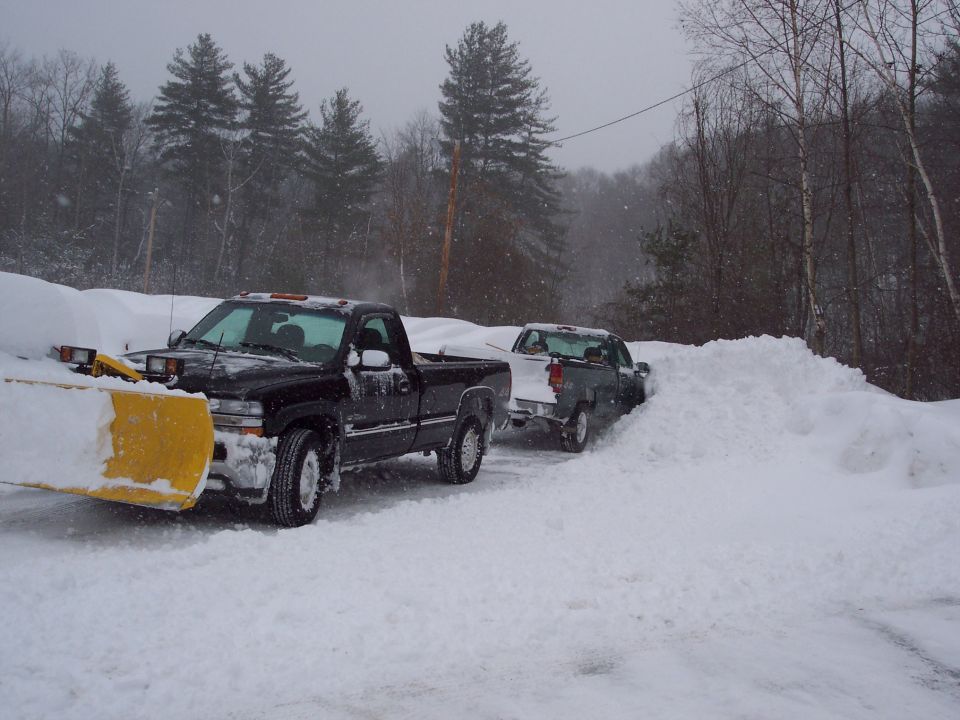 Black pickup truck with snow plow in deep snow, another truck behind