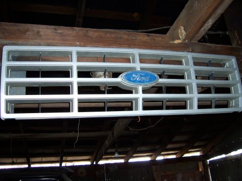 1983 Ford Bronco - A Ford grille with its iconic blue oval badge is displayed indoors, suspended from ceiling beams.