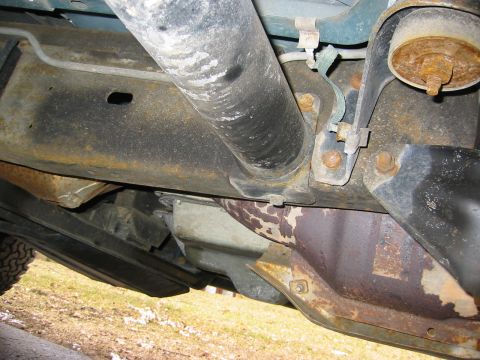 1996 Ford Bronco - Underbody view of a truck's rusty frame rail with a body-mount bushing and bolt at right, brake-line clips and brackets, a large exhaust pi…