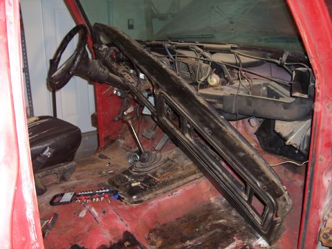 1978 Ford Bronco - Interior teardown shot through the open driver's door of a red truck, with the dash pad pulled loose and hanging forward to expose the wiri…