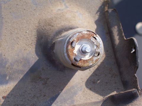 1978 Ford Bronco - A close-up view of a rusty metal bracket with a bolt and circular mounting point, likely a vehicle part.