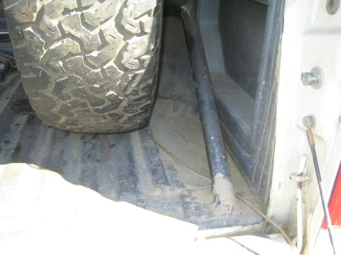 1990 Ford Bronco - chunky off-road tire rests in a dusty pickup bed next to a black bar and tie-down hardware.