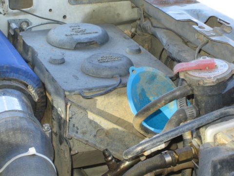 1990 Ford Bronco - dusty stamped metal panel with round caps holds a blue funnel beside hoses and a radiator cap in an engine bay.