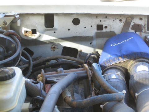 1990 Ford Bronco - engine bay with a blue silicone intake hose, a fluid reservoir and worn wiring along a white painted firewall.