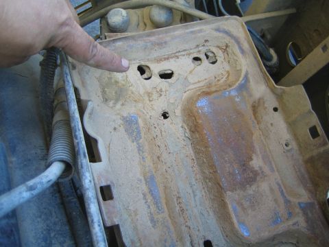 1990 Ford Bronco - hand points to drilled mounting holes on a rusty, dirt-caked stamped metal tray inside an engine bay.