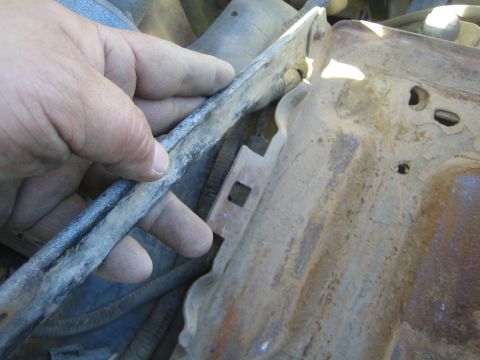 1990 Ford Bronco - grease-stained hand holds a metal rod alongside a rusty stamped bracket among engine bay components.