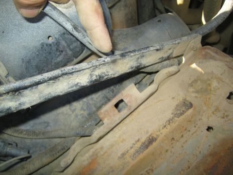 1990 Ford Bronco - finger points along a worn metal bar mounted above a rusty slotted bracket in a dusty engine bay.