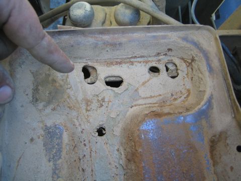 1990 Ford Bronco - finger points to corroded mounting holes in a rusty, paint-flaked stamped metal tray in an engine bay.