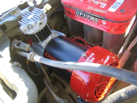 1990 Ford Bronco - red and black on-board air compressor mounted next to a red Optima battery and braided line in an engine bay.