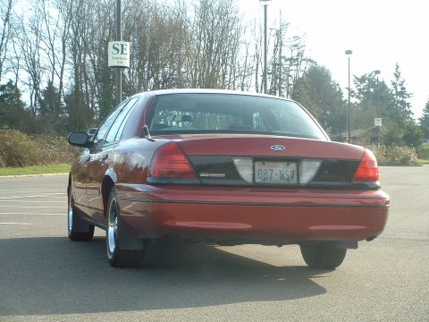 1999 Ford Crown Victoria - Rear view of a red Ford Crown Victoria parked in a lot, with trees and a sign in the background.