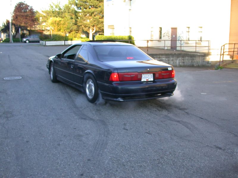 Dark blue Ford Thunderbird coupe doing a burnout with tire smoke, seen from rear three-quarter angle