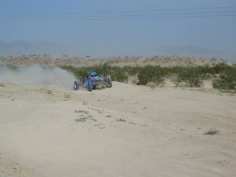 Blue off-road buggy cresting a sandy desert berm in a dust cloud