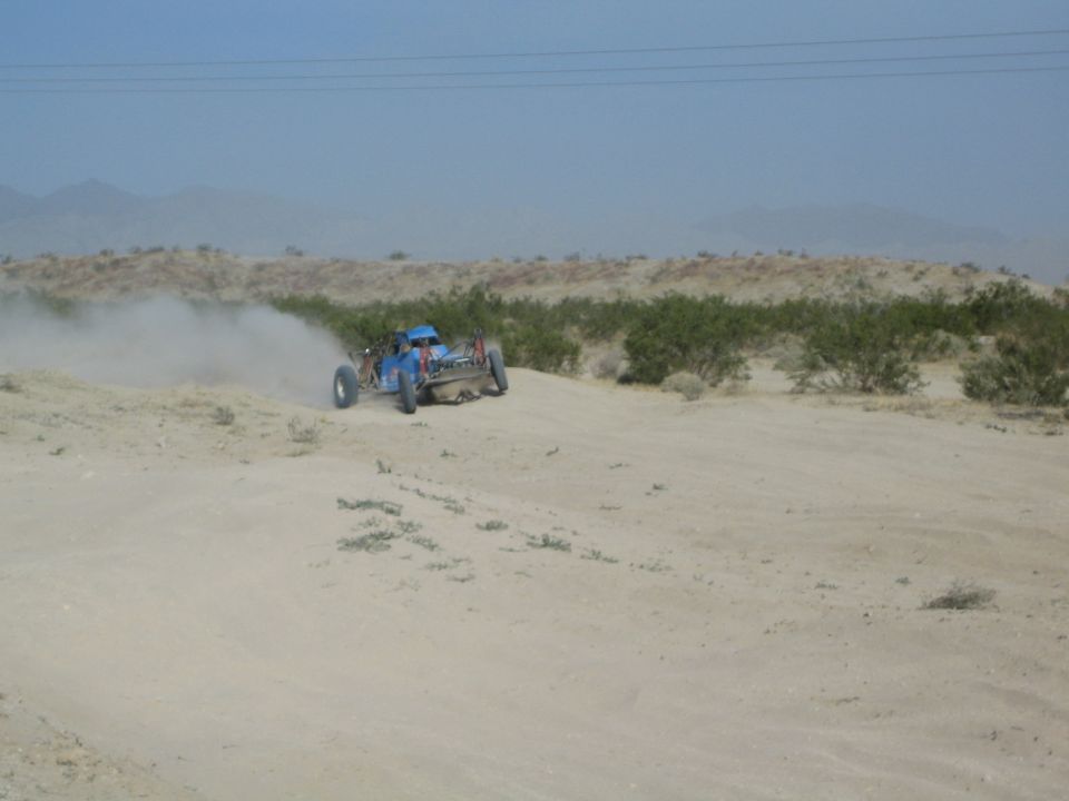 Blue off-road buggy cresting a sandy desert berm in a dust cloud