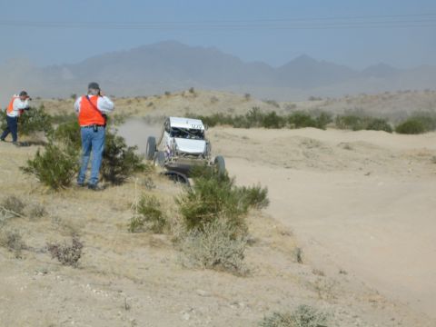 Spectators in orange vests photograph a buggy on a dusty desert course