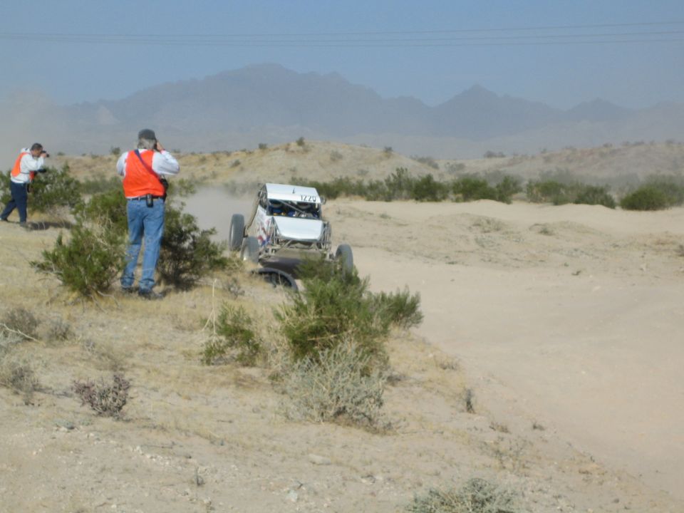 Spectators in orange vests photograph a buggy on a dusty desert course