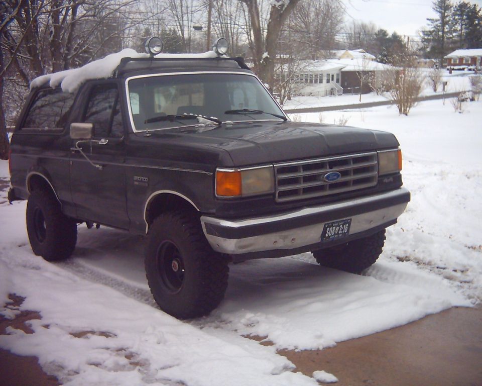 1987 Ford Bronco parked in snow, front 3/4 view. Has roof rack with lights and aftermarket wheels