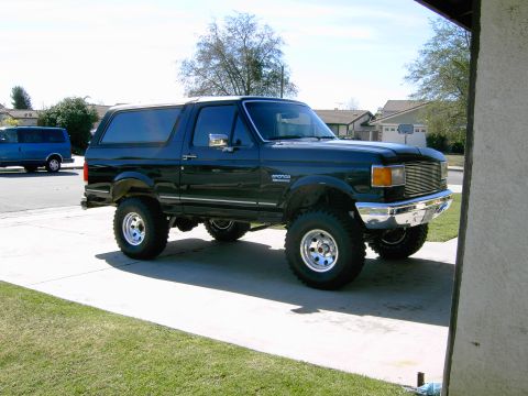 1988 Ford Bronco - A lifted dark green Ford Bronco is parked on a driveway with a blue van in the background.