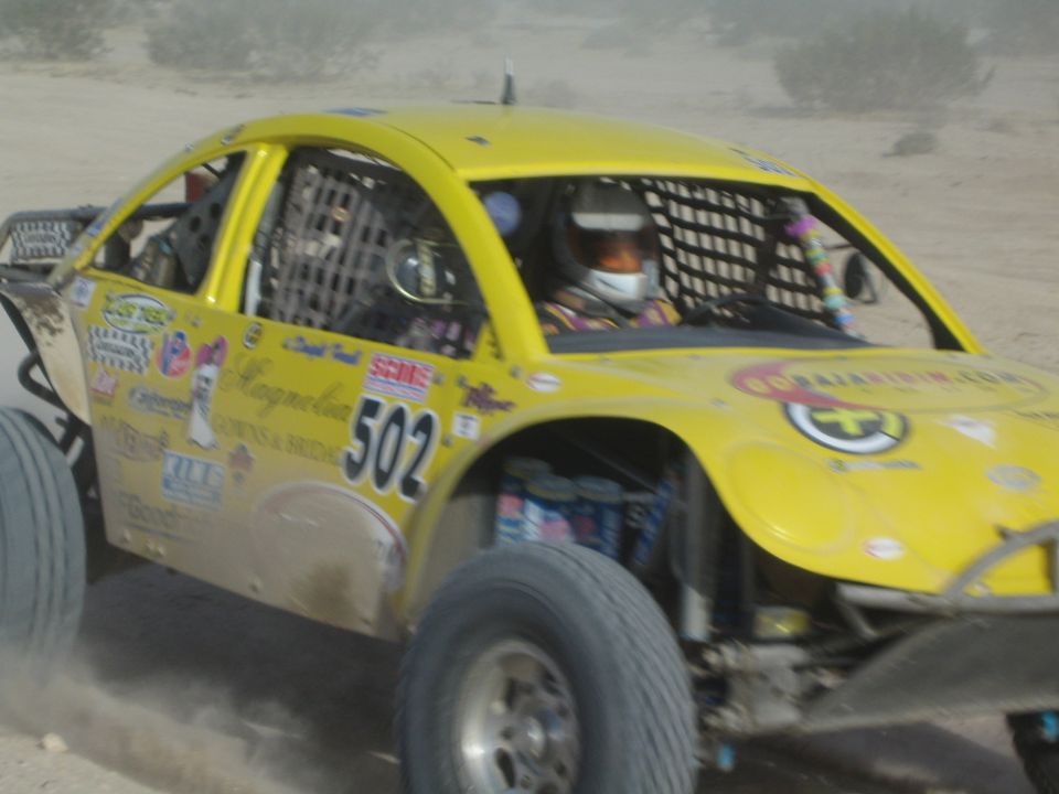 Yellow number 502 off-road race car with caged cockpit and helmeted driver