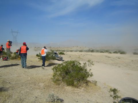 Spectators in orange vests watch a buggy near power-line towers in dusty desert