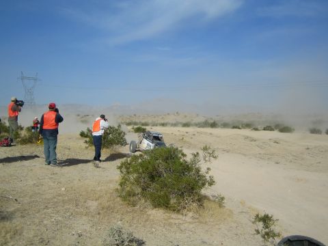 Group of vested spectators photograph a white off-road buggy in the desert