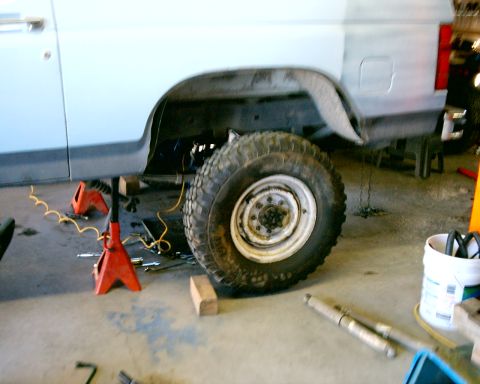 1985 Ford Bronco - A light blue Ford Bronco is elevated on jack stands for maintenance, revealing its rear wheel and suspension components.
