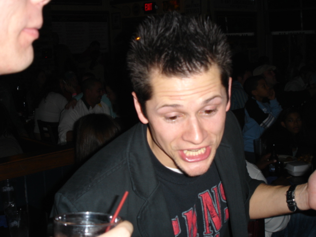 People in a dimly lit bar, one man in the foreground with spiky hair and a black jacket