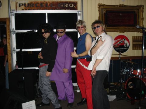 1985 Ford Crown Victoria LTD - Four men in costumes pose for a photo in what appears to be a bar or venue.