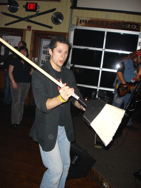 1985 Ford Crown Victoria LTD - A man holds a broom in front of a band performing on stage.