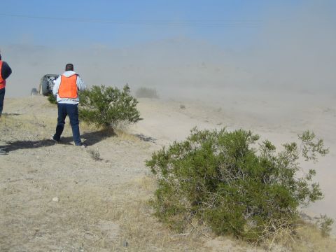 Spectators in orange vests watch a distant buggy in heavy desert dust