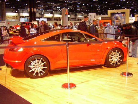 1997 Ford Taurus - An orange sports coupe is displayed at an auto show with people observing.