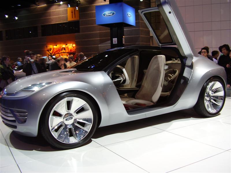 Silver concept car with butterfly doors open, showing interior and chrome wheels. Ford logo visible on display