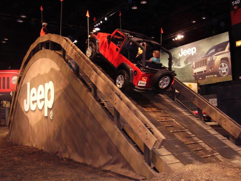 Red Jeep Wrangler driving up a wooden ramp with "Jeep" logo on the side. A person is in the driver's seat