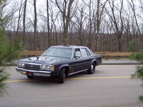 2001 Ford Crown Victoria - A dark blue sedan is parked on an asphalt lot with a wooded area in the background.