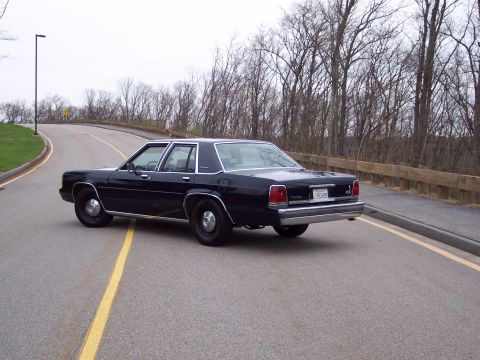 2001 Ford Crown Victoria - This black 2001 Ford Crown Victoria sedan is seen from a 3q-front angle on a road, with a wooden fence and bare trees in the background.