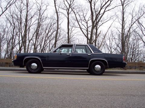 2001 Ford Crown Victoria - A dark blue 2001 Ford Crown Victoria is seen from the side, parked along a road with bare trees in the background.
