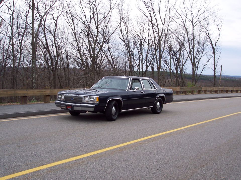 Dark blue Ford Crown Victoria parked on the side of a road with trees in the background