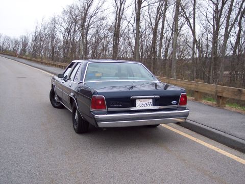 2001 Ford Crown Victoria - A dark blue 2001 Ford Crown Victoria sedan is parked on the side of a road with trees in the background.
