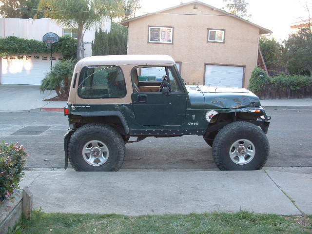 Green Jeep Wrangler parked on street, tan soft top, large tires, dented hood