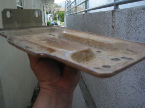 1990 Ford Bronco - rusty rectangular sheet-metal battery tray with mounting flange, held in a hand against a stucco building exterior.