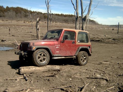1998 Jeep TJ - A red Jeep Wrangler with a tan top sits on dry, cracked earth surrounded by dead trees.